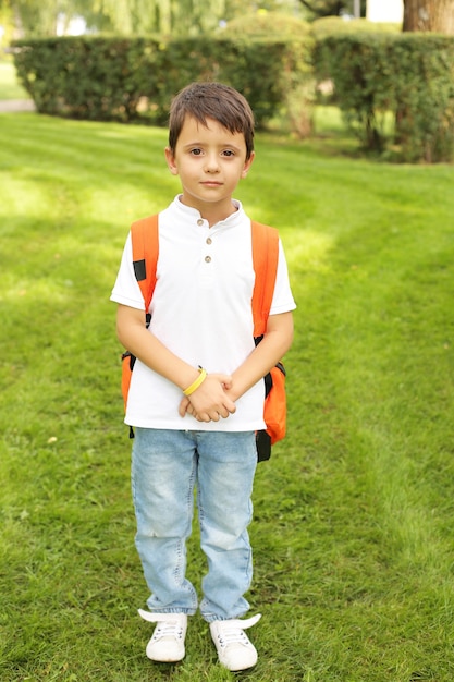 a beautiful little boy is standing in the park in a white Tshirt jeans with an orange backpack