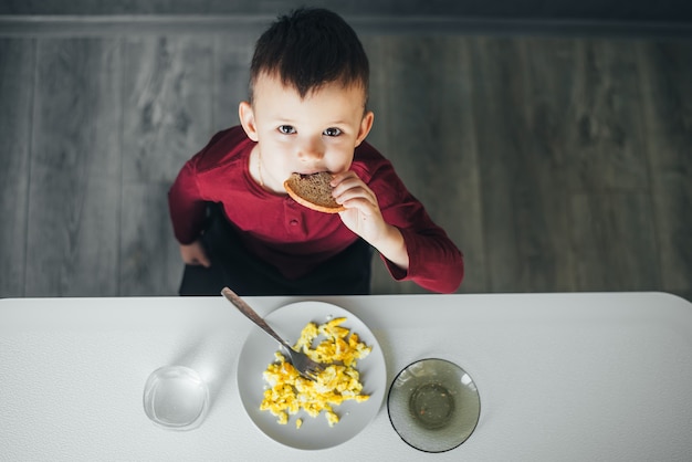 A beautiful little boy in the kitchen is eating a piece of bread and an omelet is foreshortened on top