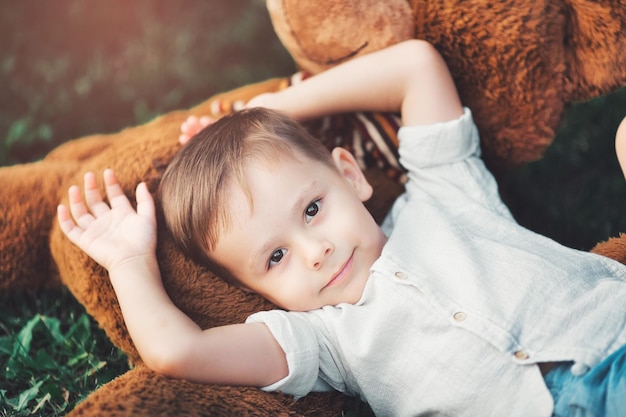 A beautiful little boy lies on the grass in the park with his favorite soft toy and looks at the camera