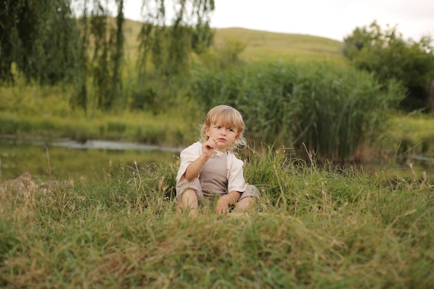 a beautiful little boy with blond hair and curly hair is resting in nature by the river