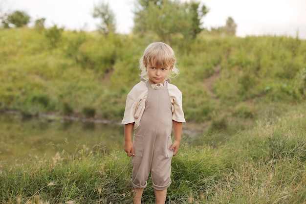 ブロンドの髪と巻き毛の美しい小さな男の子が川のそばで自然の中で休んでいます