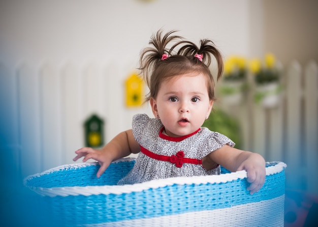 Bella bambina dagli occhi marroni si siede in un grande cestino per i giocattoli nella sua accogliente camera dei bambini.