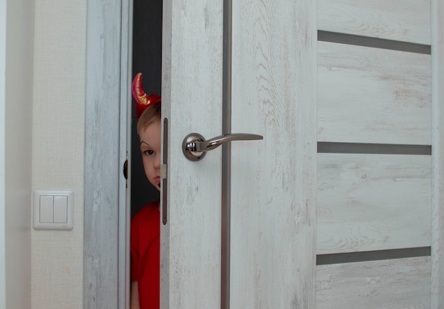 A beautiful little devil with horns. Kid. The concept of Halloween, holiday and childhood. A funny child in a carnival costume looks out from behind the door of the house. Selective focu.