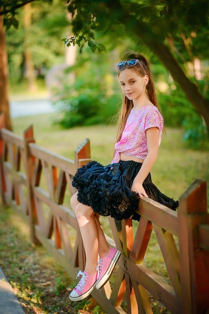 Beautiful little girl in a black magnificent tutu skirt sits on a wooden fence among green trees.