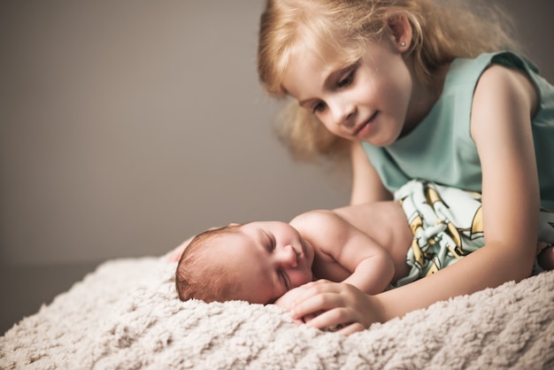 Beautiful little girl in a blue dress looks at her little charming sleeping newborn baby brother lying on a plaid on a gray wall. Family replenishment concept. Copyspace