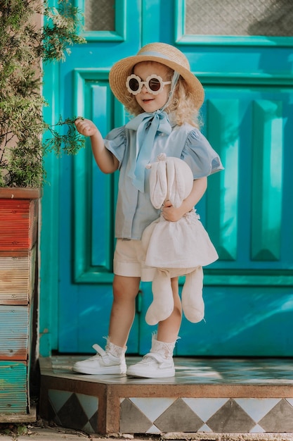 Bella bambina in abito blu, cappello di paglia e occhiali da sole che giocano all'aperto, sfondo blu, carta
