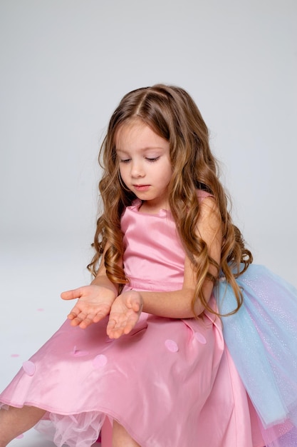 Photo beautiful little girl in a chic pink dress poses sitting on a gray background and throws confetti