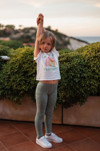 Premium Photo  Beautiful little girl child in fashionable summer clothes  with a white tshirt green leggings and white sneakers stands near a green  bush on the seashore