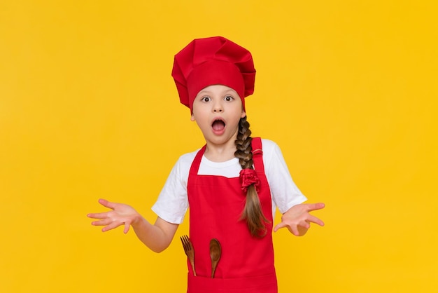 A beautiful little girl cook in a kitchen costume very much surprised A little girl chef on an isolated background the concept of how children cook food at home