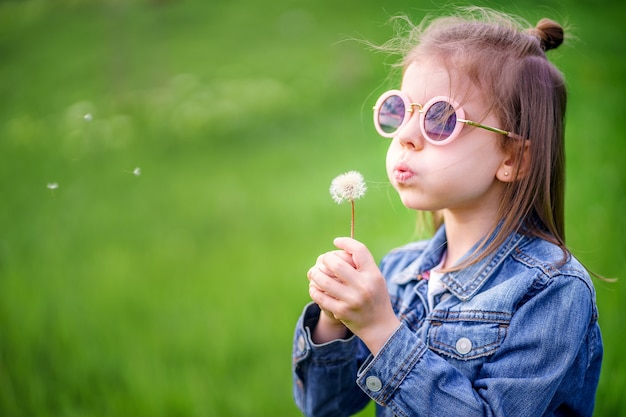 公園の屋外のタンポポに吹くデニムの服と丸いサングラスの美しい少女