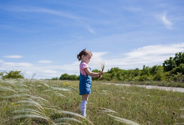 Photo a beautiful little girl in a denim sundress stands sideways in a clearing and collects a bouquet of stipa