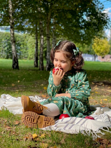 ドレスを着た美しい少女が秋の庭の綿の格子縞の上に座り、赤いリンゴを食べる