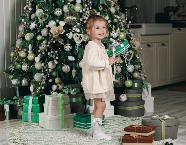 Beautiful little girl in a dress with a gift in her hand stay near the Christmas tree