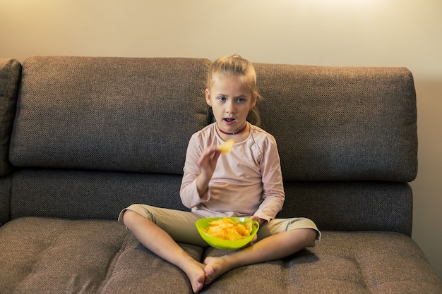 Foto bella bambina che mangia cibo malsano mentre guarda la tv sul divano