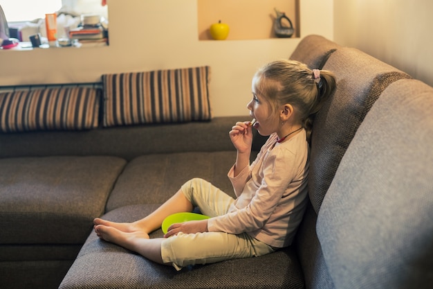 Foto bella bambina che mangia cibo malsano mentre guarda la tv sul divano