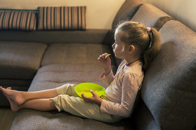 Beautiful little girl eating unhealthy food while watching tv at the sofa