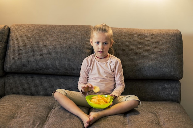 Foto bella bambina che mangia cibo malsano mentre guarda la tv sul divano