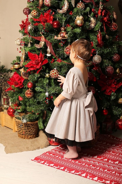 beautiful little girl in a festive dress stands by the christmas tree