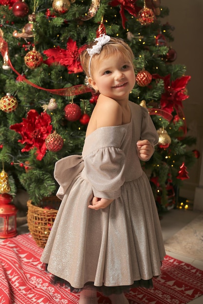 beautiful little girl in a festive dress stands by the christmas tree