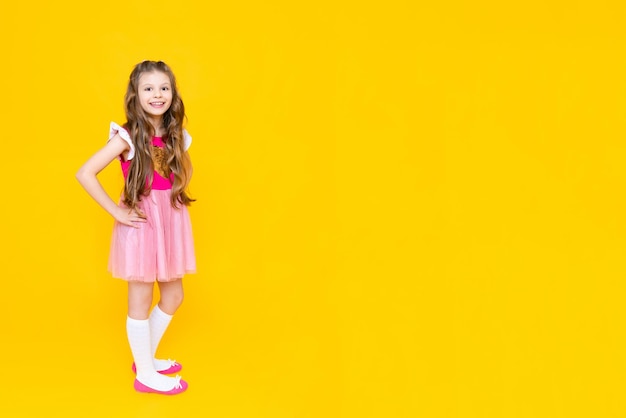 A beautiful little girl in full height in a pink dress on a yellow isolated background happy little princess with long curly hair