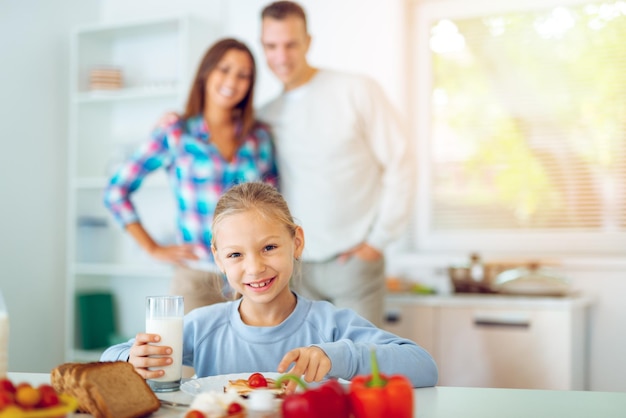 Beautiful little girl having breakfast in the domestic kitchen. Her happy parents standing behind. Looking at camera.