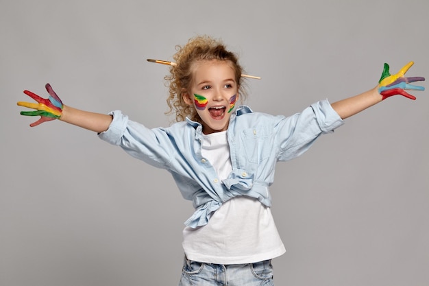 Beautiful little girl having a brush in her chic curly blond hair, wearing in a blue shirt and white t-shirt. She spread her arms widely and opened her mouth, looking away on a gray background.