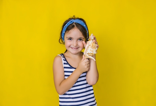 Una bella bambina che tiene una conchiglia su un muro giallo. vacanze estive.