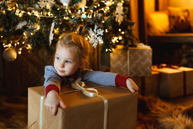 A beautiful little girl hugs a box with a Christmas gift in front of a decorated Christmas tree. 