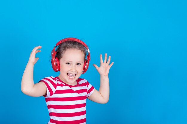 A beautiful little girl is enthusiastically listening to music with red headphones on blue background