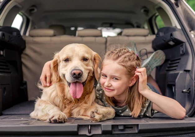 Beautiful little girl lying with golden retriever dog in the\
car trunk and looking back. child kid with purebred doggy pet with\
tonque out in the vehicle at the nature together