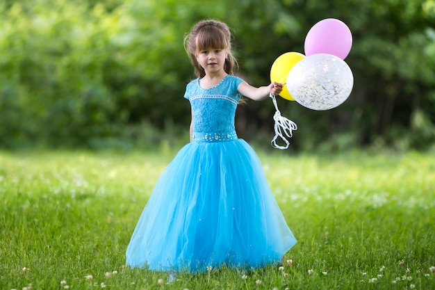 Beautiful little girl in nice long blue evening dress looking like princess looks  holding colorful balloons 