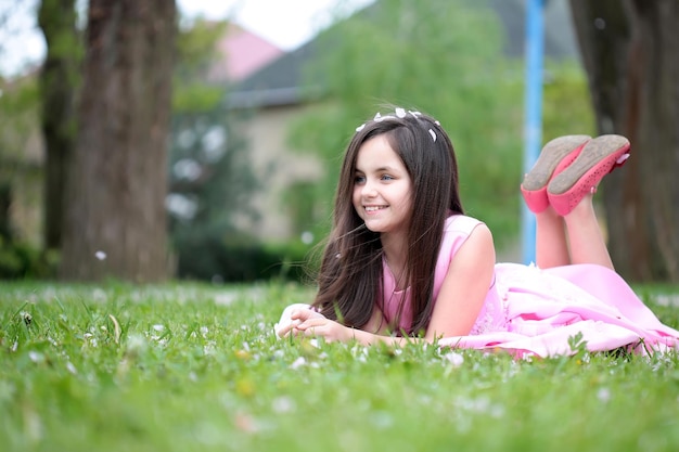 Beautiful little girl in pink dress with long brunette hair and smiling face lying on green grass in...