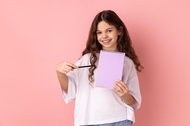 Красивая маленькая девочка, указывающая на бумажный блокнот и улыбающаяся в камеру, имеет позитивное выражение лица