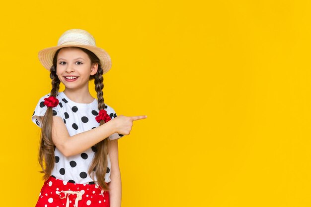 A beautiful little girl in polka dot shorts and a straw hat is pointing at your advertisement Summer holidays A teenage girl is waiting for summer on a yellow isolated background Copy Space