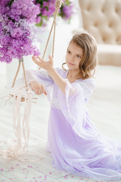 Beautiful little girl of preschool age in a delicate dress sitting on a swing decorated with flowers of lilac