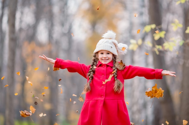 Beautiful little girl in red coat with autumn leaves outdoors in a park.