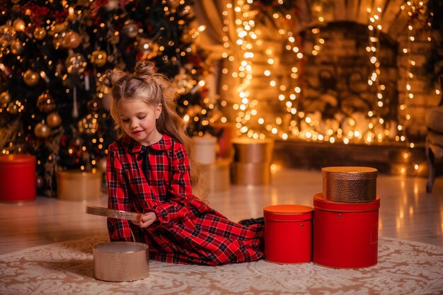 Beautiful little girl in a red dress at home near the tree opens gifts