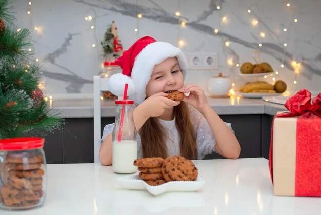 サンタの帽子をかぶった美しい少女は、家でクリスマスイブにミルクを飲み、クッキーを食べます。休日、クリスマス、子供時代の概念。セレクティブフォーカス