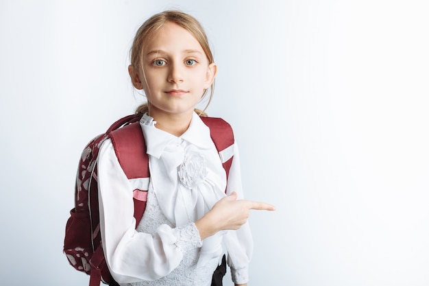 Una bella studentessa bambina con valigetta che punta a parete, muro bianco