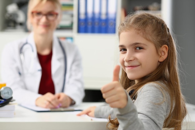 La bella bambina mostra i pollici in su e il medico si siede sullo sfondo