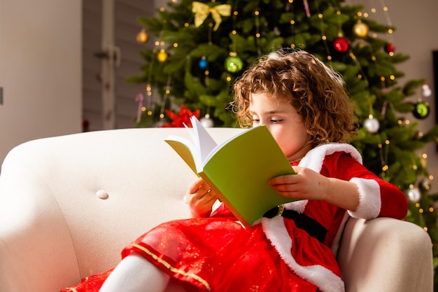 Photo beautiful little girl sits on the chair next to a nicely decorated christmas tree