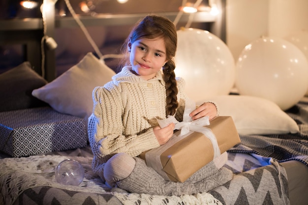 Красивая маленькая девочка сидит на кровати с подарком в руках
