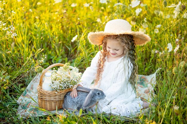 beautiful little girl sitting in a straw hat in a chamomile field with a rabbit
