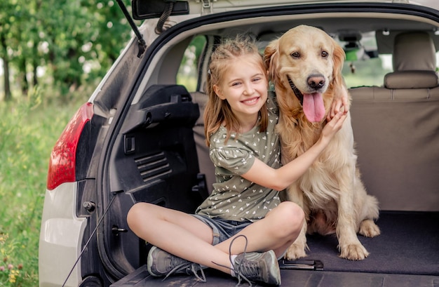 車のトランクにゴールデンレトリバー犬と一緒に座って、カメラを見て笑っている美しい少女。自然の中で車の中で純血種の犬のペットを抱き締める子供の子供