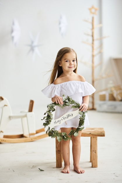 A beautiful little girl sitting on a wooden stool in a beautiful dress in the white room