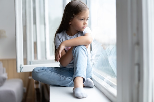 Beautiful little girl smiling and watching out the window. A child looks out the window. Young girl looking from window. Portrait of cheerful kid lies at windowsill.