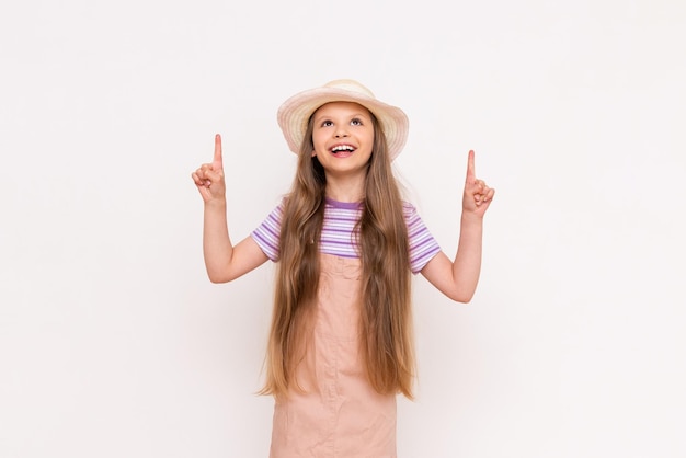 A beautiful little girl in a summer sundress and a straw hat on a white isolated background points her index finger up at your advertisement