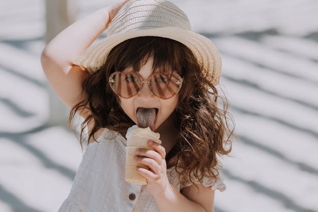 夏のビーチでアイスクリームを食べるサングラス白いドレスと帽子の美しい少女