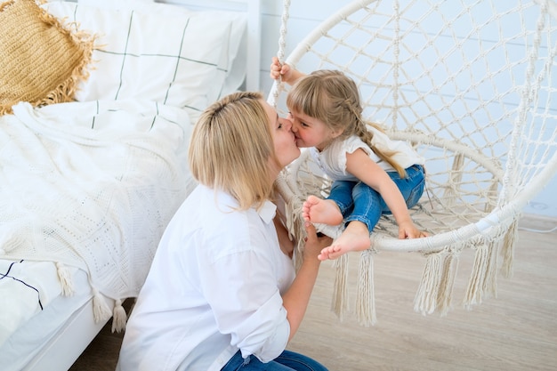 Photo beautiful little girl swinging in a hanging chair in the bedroom with her mom baby kisses mommy