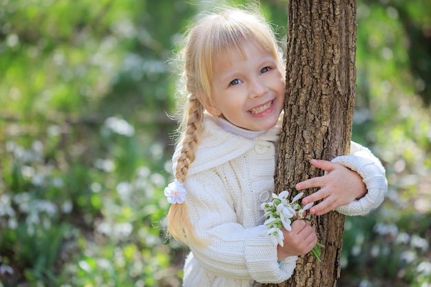 写真 スノードロップの花束を持つ美しい少女。白いニットのセーターを着た少女が木の幹を抱きしめます。明るく晴れた春の日